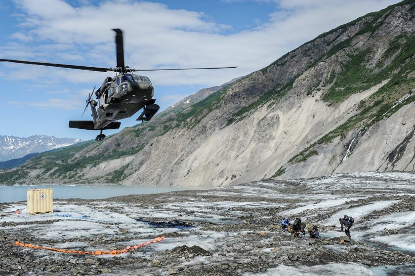 Members of a specialized investigative team from the Join POW/MIA Accounting Command wait as a UH - 60 Blackhawk from Joint Base Elmendorf-Richardson comes in for a landing to transport them back after a day of assessing a historic aircraft crash site. The mission of the Joint POW/MIA Accounting command is to conduct global search, recovey and laboratory operations to identify unaccounted-for Americans from past conflicts in order to support hte Department of Defense's personnel accounting efforts. (Clifford Bailey / US Navy)