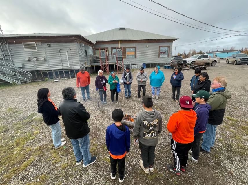 People from kids to elders stand in a circle around a fire, many of them wearing orange shirts
