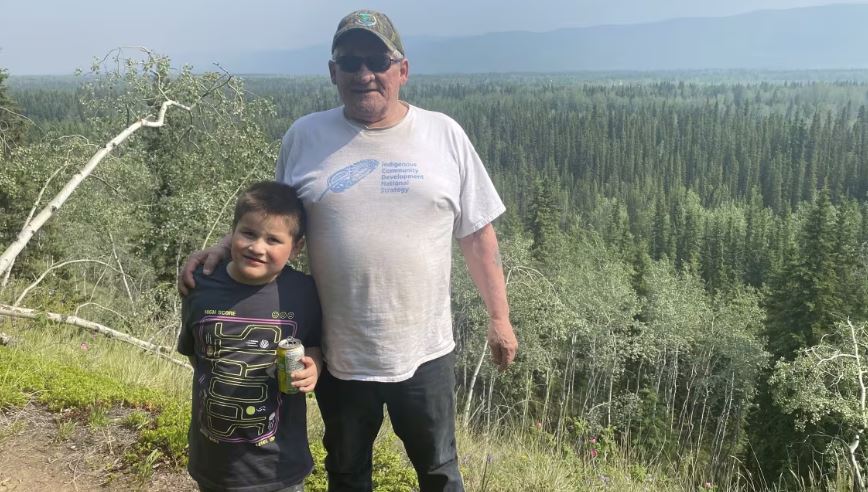 An older man stands with a young boy with forest and mountains behind them.
