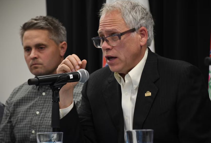 A man speaks into a microphone as another man looks on.