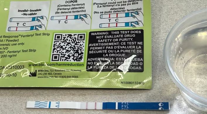 A strip of thin material with blue markings and two red lines lies on a flat surface below a package with images of a hand holding a similar strip with one or two stripes on it, and indicating which are indicators a substance has fentanyl and which does not. A small plastic container of liquid stands beside them.
