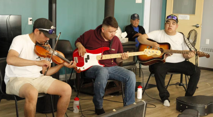 Three men seated in a room playing fiddle, guitar and bass.