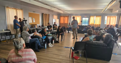 A group of people seated in a room, with a man standing at the centre with a microphone.