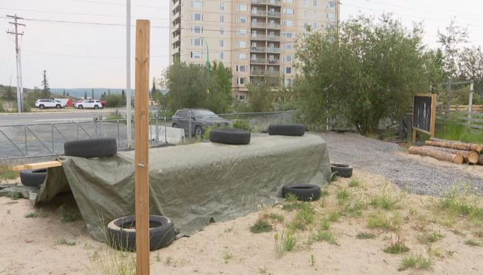 A schoolyard area under construction with logs, tires, and a structure covered by a tarp.
