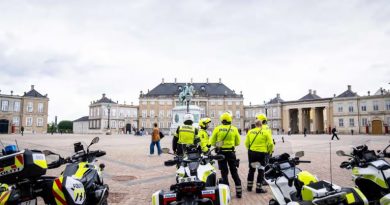 Four people wearing yellow jackets with the word POLITI stand near four motorcycles on a square surrounded by ornate old buildings.