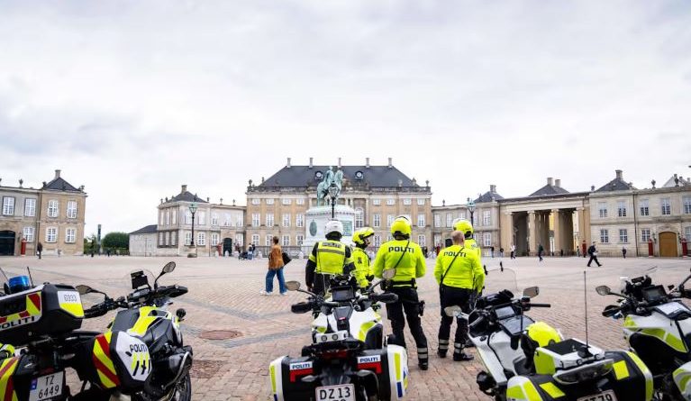 Four people wearing yellow jackets with the word POLITI stand near four motorcycles on a square surrounded by ornate old buildings.