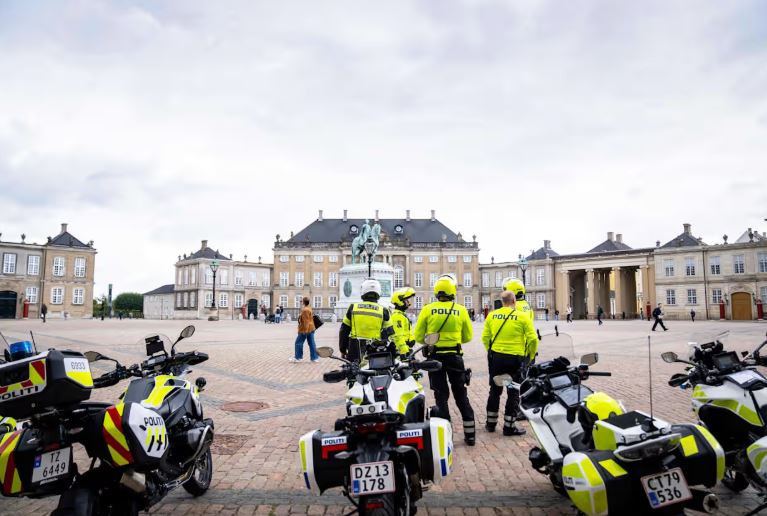 Four people wearing yellow jackets with the word POLITI stand near four motorcycles on a square surrounded by ornate old buildings.