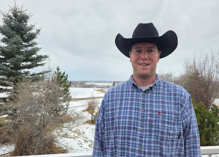 A man in a blue flannel shirt and cowboy hat standing outdoors