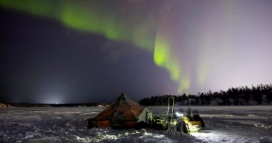 A tent on snow-covered barren land with northern lights dancing in the sky at night.
