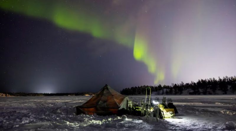 A tent on snow-covered barren land with northern lights dancing in the sky at night.