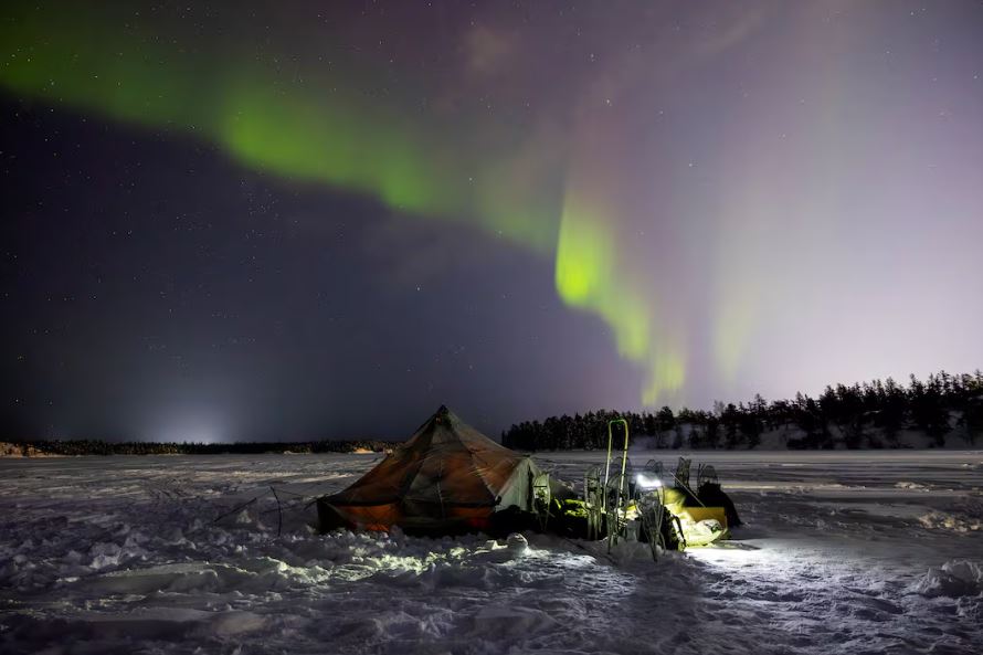 A tent on snow-covered barren land with northern lights dancing in the sky at night.