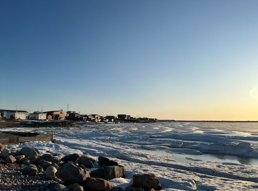 A photo of a shoreline with sea ice and some small houses in the background 