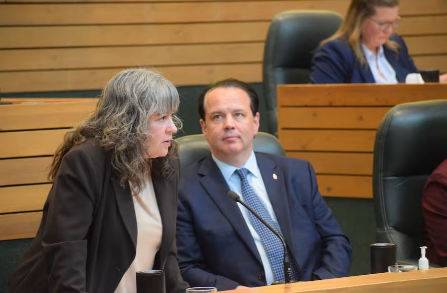 Woman speaks into microphone as man behind her looks off in distance in the Yukon legislature.