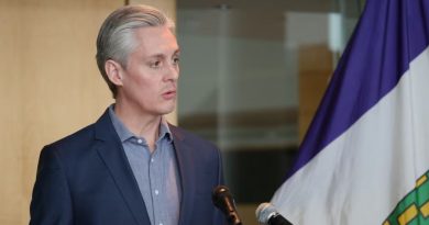 A man in a suit stands before 2 microphones, speaking.
