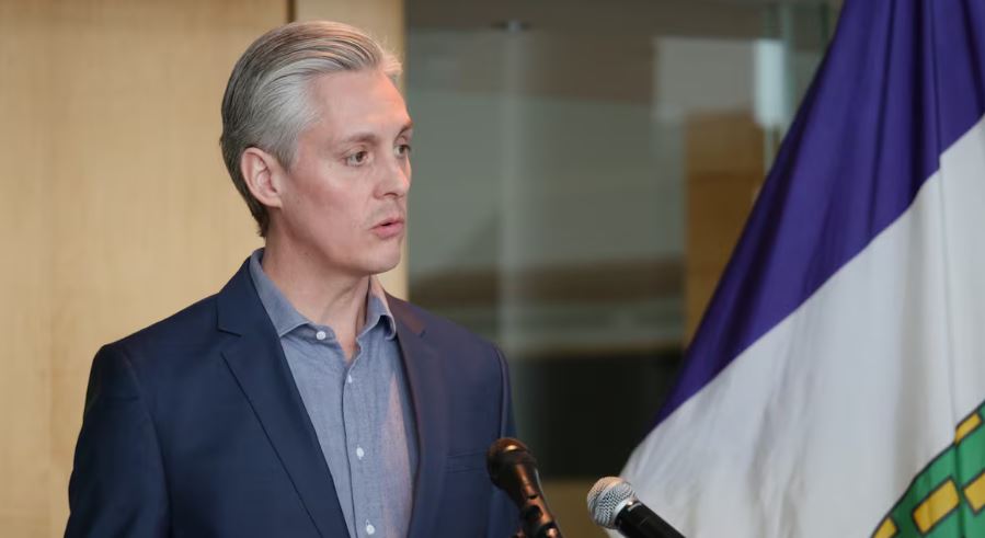 A man in a suit stands before 2 microphones, speaking.