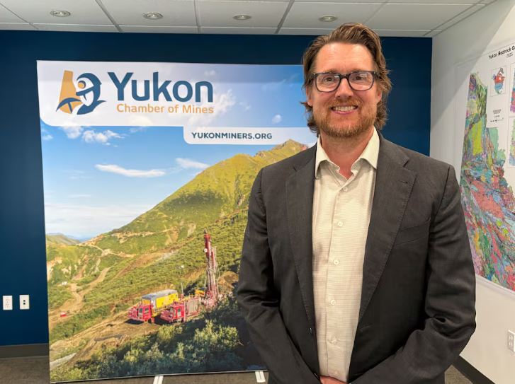 A man with brown hair wearing a black blazer, white shirt and glasses standing in front of a sign that says Yukon Chamber of Mines.