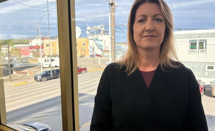 A woman with dark blonde hair wearing a black sweater standing in front of a window overlooking a city.