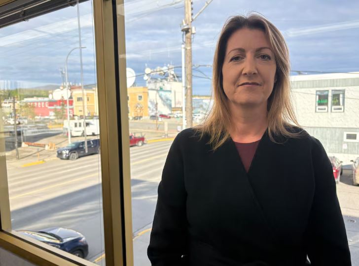 A woman with dark blonde hair wearing a black sweater standing in front of a window overlooking a city.
