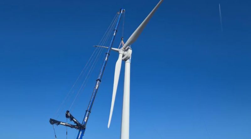 A blue crane lifting blades on a white turbine against a blue sky.