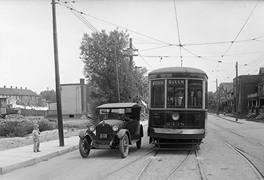 Dans les années 1920, à Toronto, une automobile et un tramway partagent la voie à l’angle des rues Queen et Bay. (Archives publiques, Ville de Toronto)