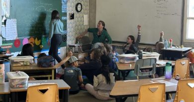 Une enseignante écrit au tableau tandis que des élèves sont assis par terre autour d'elle.