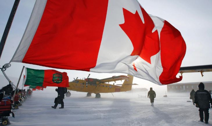 Une photo montre un drapeau canadien qui flotte lors d'une patrouille de souveraineté sur l'île d'Ellesmere, au Nunavut. On y voit des membres des forces armées accompagnés d'un groupe de réservistes.