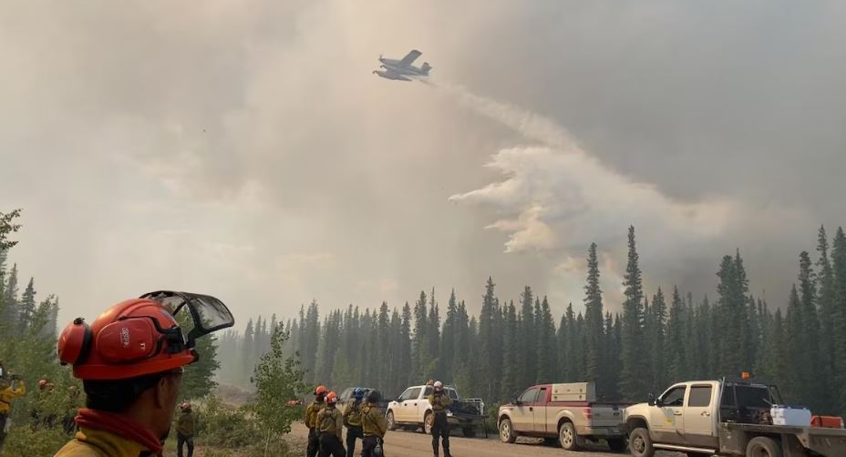Avion-citerne qui déverse de l'eau sur la forêt pendant que des pompiers forestiers sont au sol et observent la scène, en juin 2024, à Fort Good Hope, aux Territoires du Nord-Ouest.