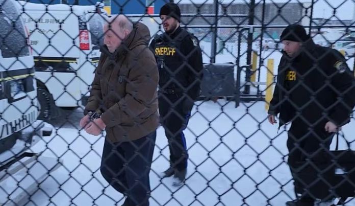 A man is seen through a fence walking in handcuffs with two sheriffs behind him