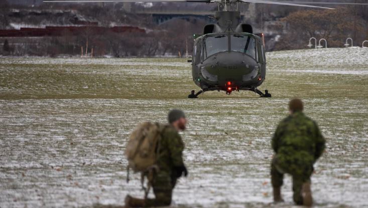 Deux membres militaire accroupis avec au loin l'hélicoptère CH-146 Griffon de l'Aviation royale canadienne.