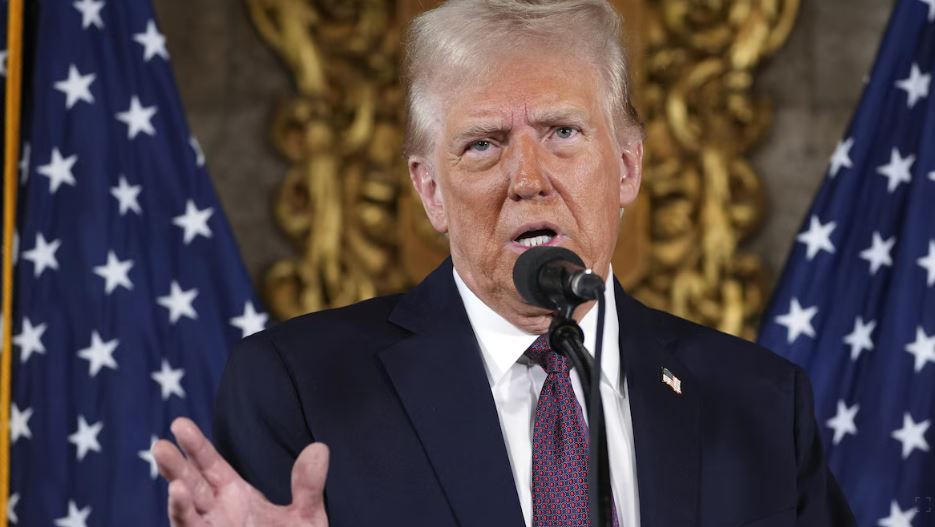 Donald Trump au micro, devant des drapeaux américains, bouge la main en parlant.