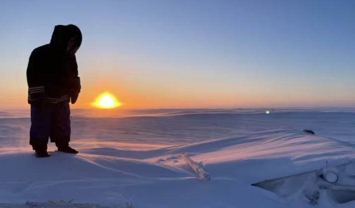 Une photo montrant un personne se tenant au sommet d'une hutte de terre recouverte d'une épaisse couche de neige, le 18 janvier 2025, au Nunavut.