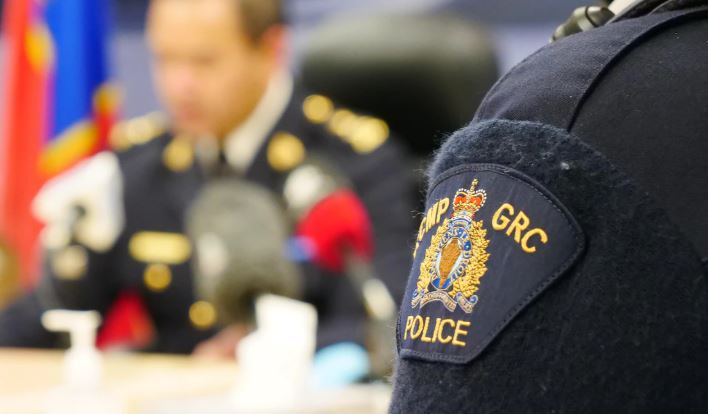 Gros plan sur le blason de la GRC cousu sur un chandail d'un membre de la force. Au deuxième plan, flou, un agent de police s'exprime aux micros.