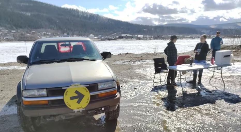 Un bureau de vote installé en extérieur au bord du fleuve Yukon à côté d'une camionnette et trois personnes.