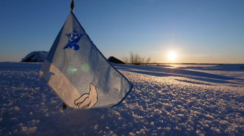Un petit drapeau bleu et blanc arborant les symboles d'un ours polaire et d'un logo représentant une moitié de fleur de lys et une moitié de flocon est planté dans la neige.