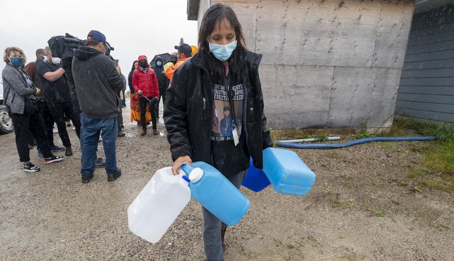 Sous une pluie battante, une femme marche, le visage couvert d'un masque médical pendant la pandémie, en transportant quatre bidons de plastique.