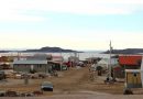 Une rue en terre à Kugaaruk bordée de bâtisses basses et de plusieurs voitures et quads.