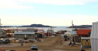 Une rue en terre à Kugaaruk bordée de bâtisses basses et de plusieurs voitures et quads.
