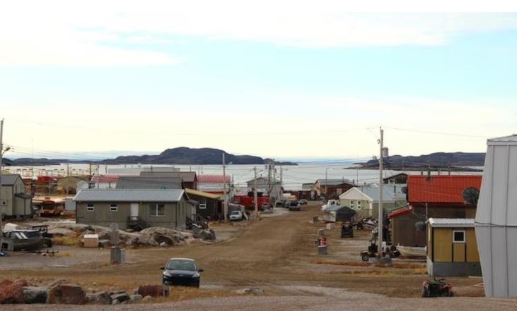 Une rue en terre à Kugaaruk bordée de bâtisses basses et de plusieurs voitures et quads.