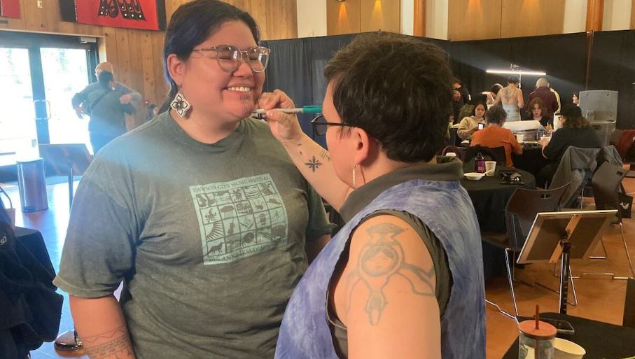 Une femme sourit pendant qu'une artiste autochtone applique, avec précision, un tatouage traditionnel sur son menton au sein d’un espace communautaire, rempli de participantes engagées dans la redécouverte de leurs pratiques ancestrales.