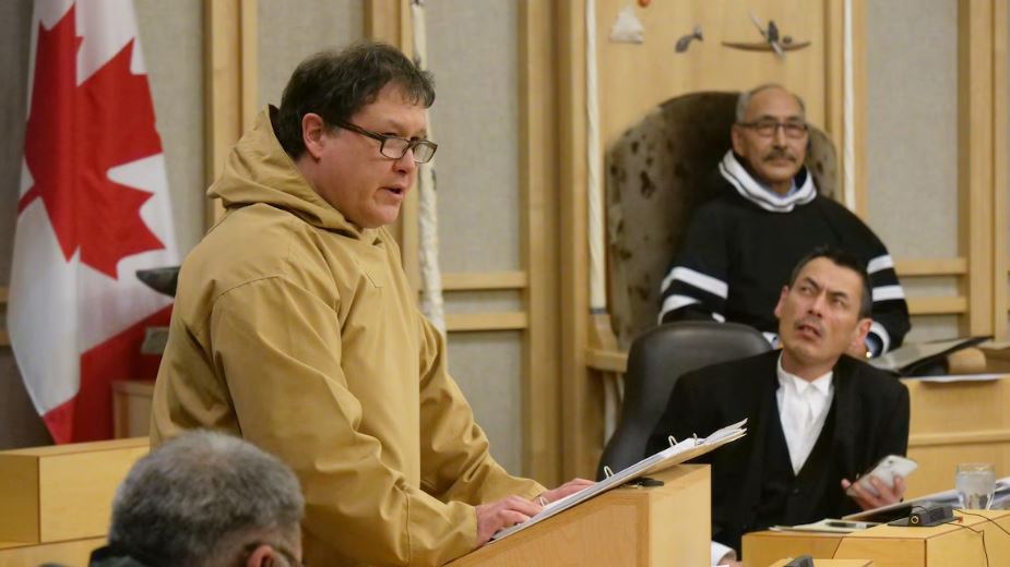 George Hickes debout à l'Assemblée législative entouré de ses collègues.