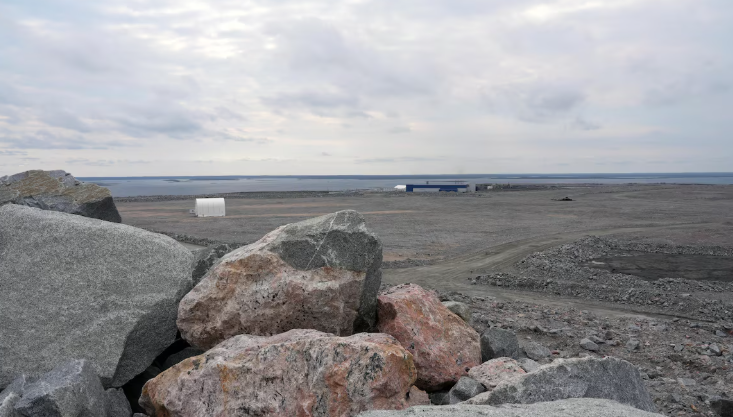 De grosses roches empilées, avec en arrière-plan un bâtiment de la mine et un plan d'eau, en août 2024.