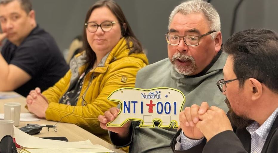 Un homme assis à côté de plusieurs personnes tient une plaque d'immatriculation en forme d'ours polaire, à Iqaluit, au Nunavut, le 20 mai 2025.
