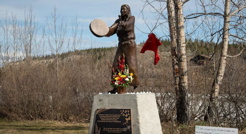 Une statue en bronze représentant une femme en train de faire du tambour, un bouquet de fleurs est posé sur son socle, à Whitehorse.
