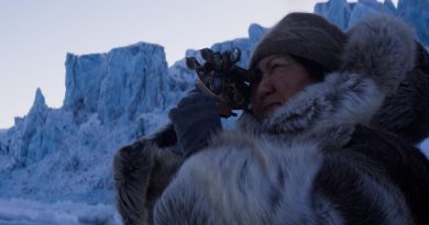 Une photographie tirée d'une scène de film montrant une femme vêtue d'un manteau en peau de caribou tient des jumelles antiques dans ses mains.