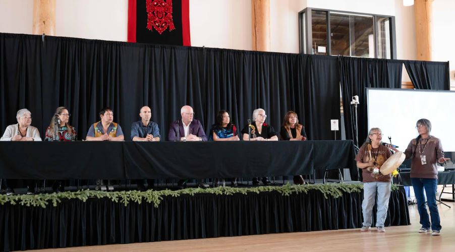 Huit personnes assises à une longue table et deux autres debout qui tiennent un tambour traditionnel.