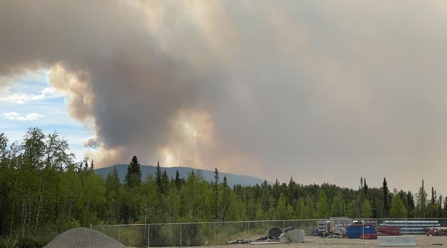 Une épaisse fumée s'élève au-dessus d'une montagne avec en avant-plan du matériel de construction gardé par une clôture.