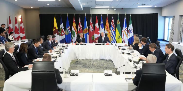 Les premiers ministres du Canada assis autour d'une table blanche, les drapeaux du Canada, des provinces et des territoires en arrière-plan.