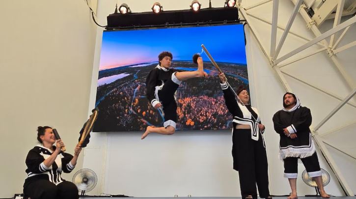 Un jeune homme saute pour toucher un bâton lors d’une démonstration de jeu inuk à l’Expo d’Osaka, À gauche, une femme est assise et tient un tambour, tandis qu’une autre femme et un homme en tenue traditionnelle assistent à la scène.