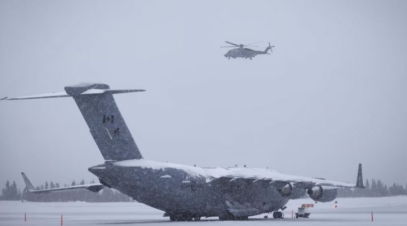 Un avion militaire canadien est à l’arrêt sur une piste enneigée tandis qu’un hélicoptère militaire vole au-dessus.