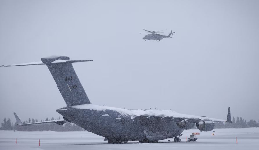 Un avion militaire canadien est à l’arrêt sur une piste enneigée tandis qu’un hélicoptère militaire vole au-dessus.